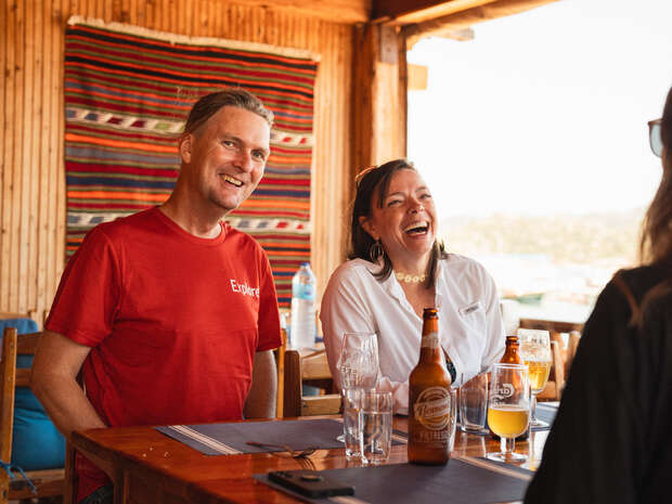 Two people are sitting at a wooden table, laughing and enjoying drinks. They are in a cozy, warmly-lit setting with a colorful tapestry on the wall behind them, adding to the relaxed atmosphere.