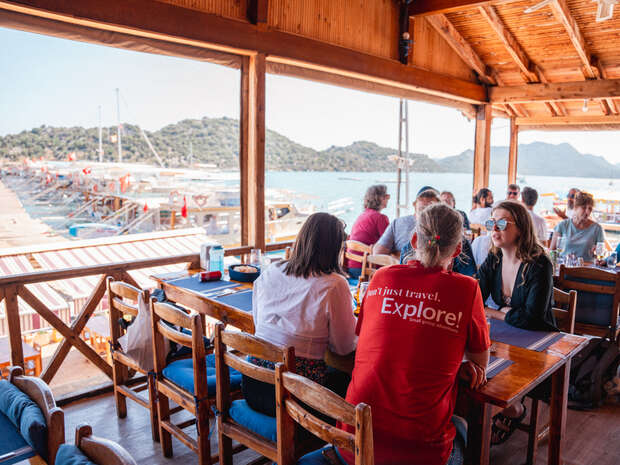 People sit at a wooden table in an open-air restaurant. One person wears a shirt reading, "Don't just travel. Explore!" Overlooks a marina with boats and distant hills.