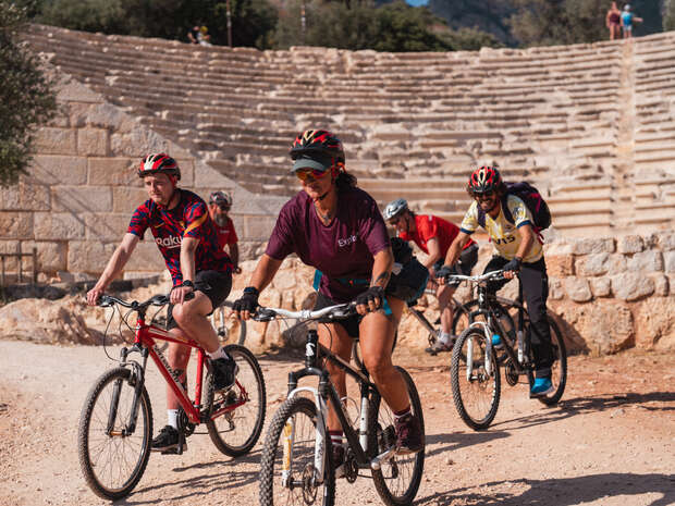 Cyclists ride on gravel paths through an ancient amphitheater, wearing helmets and casual sportswear. Sunlight illuminates the stone seating in the background, and vegetation is visible on the horizon.