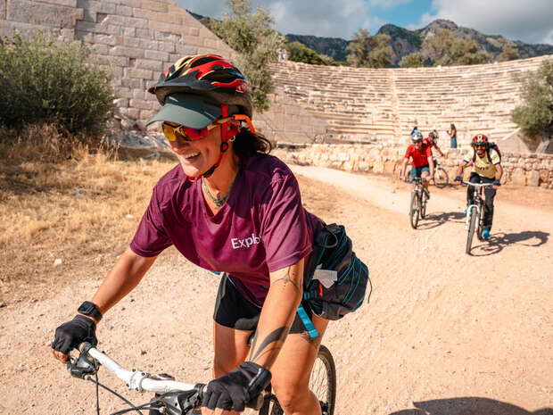 A cyclist, wearing a helmet and "Explore" shirt, rides energetically on a dirt path beside ancient stone ruins, followed by two other cyclists in a sunny, mountainous landscape.