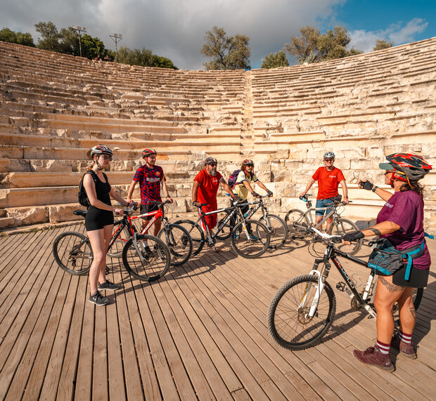 Cyclists stand with their bikes on a wooden stage of an ancient amphitheater, discussing or preparing for an activity. Stone seating surrounds them; trees and clouds visible above.