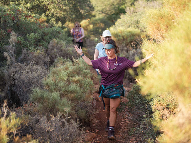 A woman in a purple "Explore" shirt walks with arms outstretched along a narrow trail, surrounded by dense, green and brown bushes. Two other people follow behind her.
