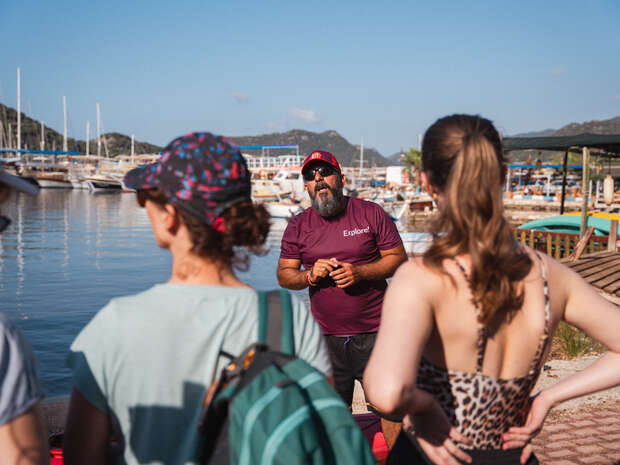 A bearded man in a maroon "Explore" shirt talks to three people near a dock, with boats and hills visible under a clear blue sky.