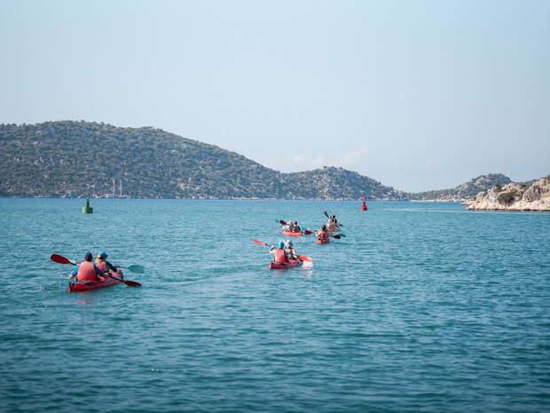 People paddle red kayaks in a line across a calm, blue sea, surrounded by distant hills and buoys, under a clear sky.