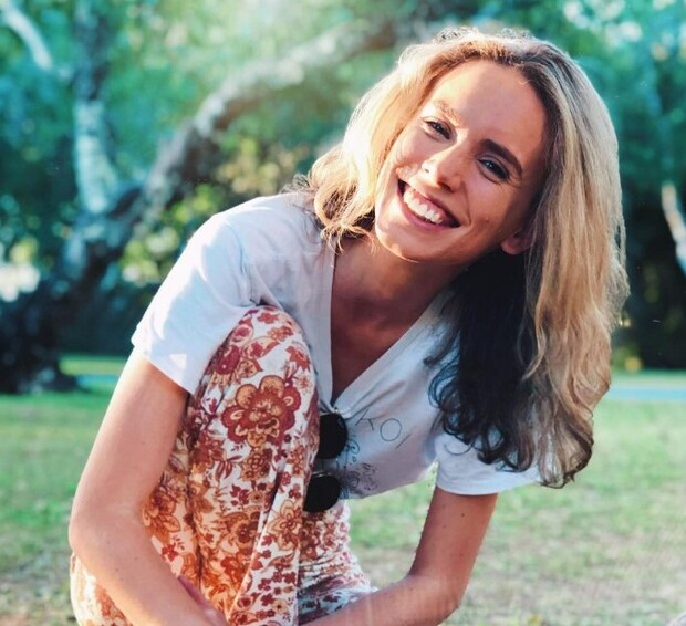 A person smiles while crouching slightly, wearing a floral patterned outfit. Bright natural light illuminates the scene, set in a grassy park with blurred green trees in the background.