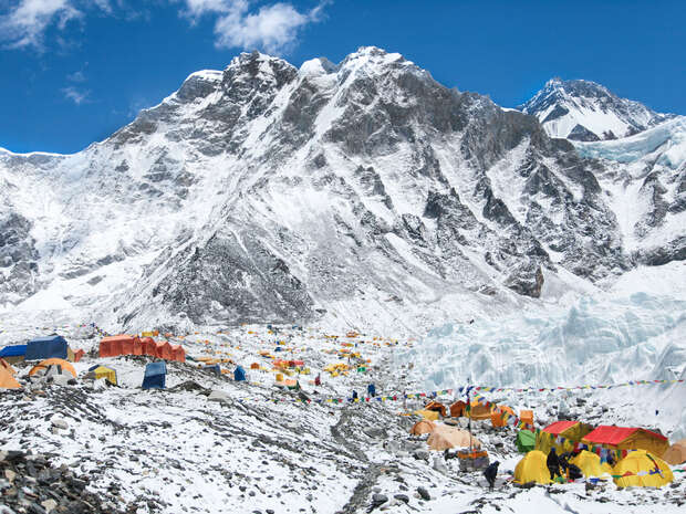 Colorful tents are pitched on a snowy landscape, forming a camp at the base of towering, snow-covered mountains under a partly cloudy sky.