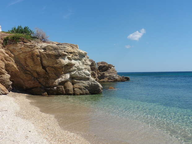 Rocky cliffs rise sharply from a secluded beach, curving into a calm, clear sea under a bright blue sky. Sparse greenery tops the cliffs, and a lone cloud floats overhead.