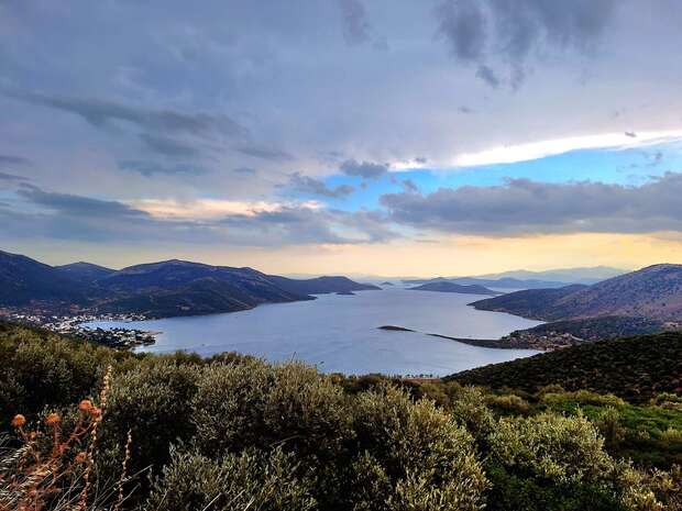 A serene bay stretches between rugged hills, under a dramatic sky with scattered clouds and patches of blue. Dense greenery covers the foreground, creating a tranquil natural landscape.