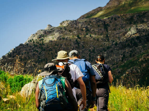 Hikers wearing backpacks walk along a grassy trail, surrounded by rocky mountains and clear skies.