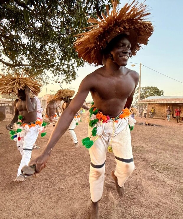 A person in a straw hat and colorful outfit dances energetically, holding objects, outdoors on a dirt surface, surrounded by others in similar attire and trees under a bright sky.