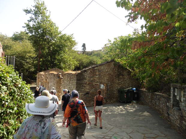 A group of people walks along a sunny stone path beside an ancient stone building, surrounded by lush green trees and scattered brown leaves, creating a serene outdoor setting.