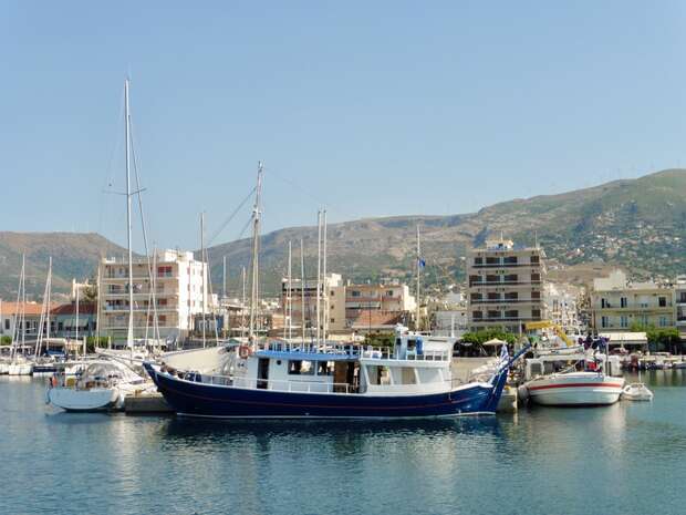 A blue fishing boat is docked at a marina, surrounded by several smaller boats. In the background, a coastal town with multi-story buildings lies beneath green hills.