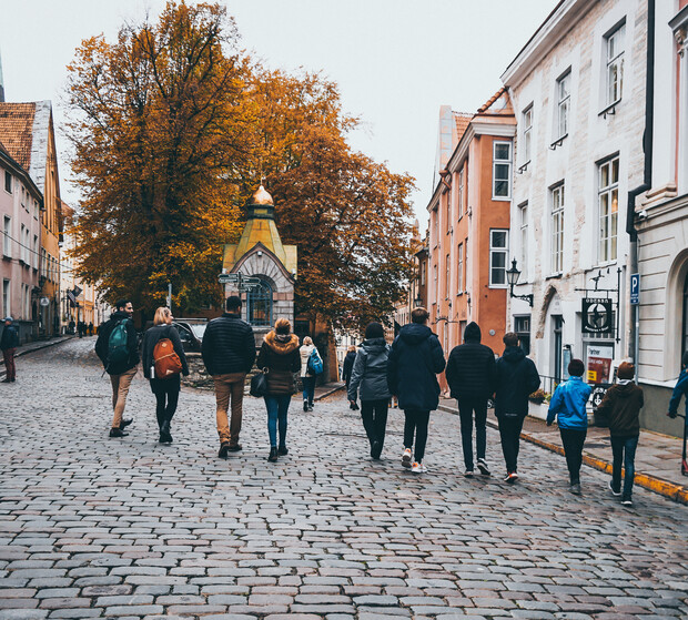 People walking on a cobblestone street, surrounded by historic buildings with autumn trees nearby. A small, ornate structure is visible ahead, evoking a quaint, old-town atmosphere.