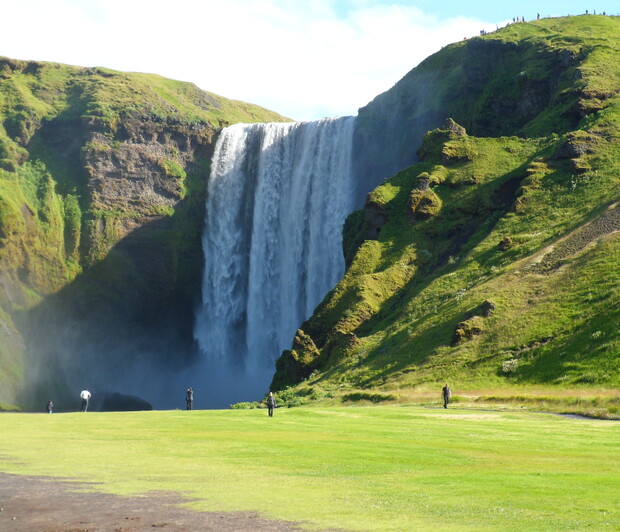 A waterfall cascades down a rocky cliff surrounded by lush green hills. People stand on a grassy field below, observing the scene under a clear blue sky.