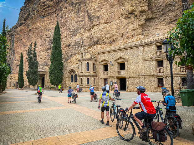 Cyclists ride through a cobblestone courtyard surrounded by tall, rocky cliffs and ancient stone buildings. Large trees and lampposts line the path, creating a scenic, historical atmosphere.