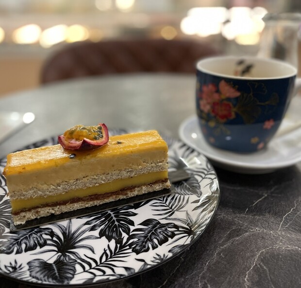 A slice of layered passion fruit cake rests on a leafy-patterned plate, beside a blue floral teacup on a marble table, in a softly lit cafe setting.