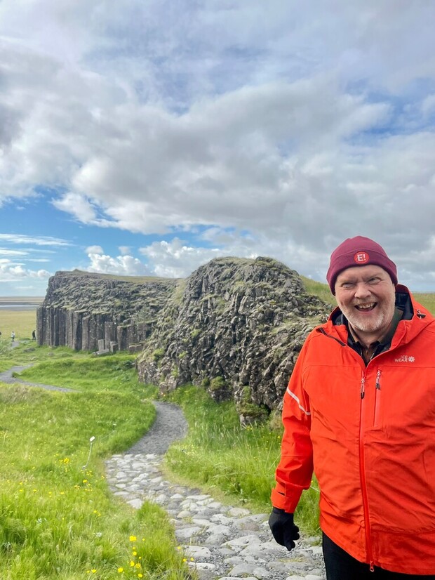 A person in a red jacket and hat stands smiling on a cobblestone path. Behind them, there are large rock formations in a grassy landscape under a cloudy sky.