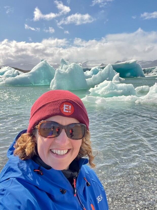 A person wearing a blue jacket and red beanie smiles at the camera. They are standing near a body of water with large icebergs in the background under a partly cloudy sky.