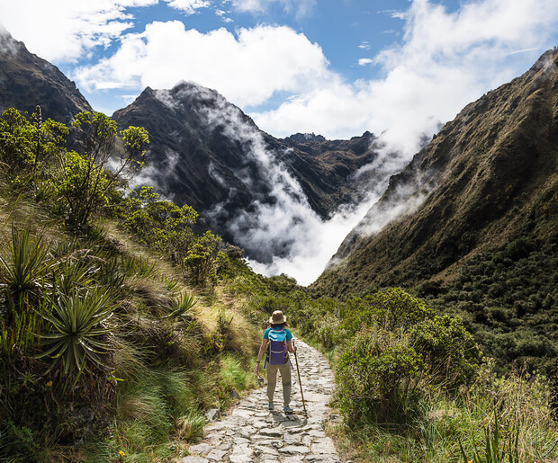 A lone hiker walks along a stone path, surrounded by lush greenery and mountainous terrain, with clouds swirling around the peaks in the background.