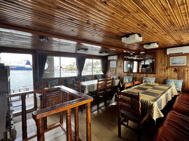Wood-panelled dining area featuring long tables with checkered tablecloths and chairs; windows reveal a waterfront view, creating a cozy, nautical ambiance on a docked boat.