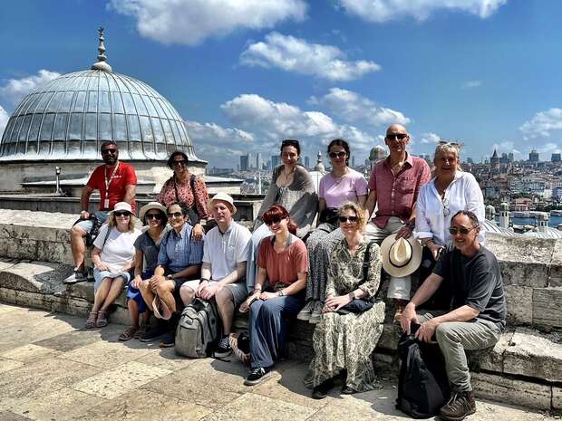 A group of thirteen people, some seated, some standing, pose on a historical rooftop under a dome beside city buildings against a partly cloudy blue sky.
