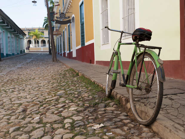 A green bicycle leans against a cobblestone street's curb beside colorful colonial buildings with bars on the windows and a hanging sign, under a clear sky.