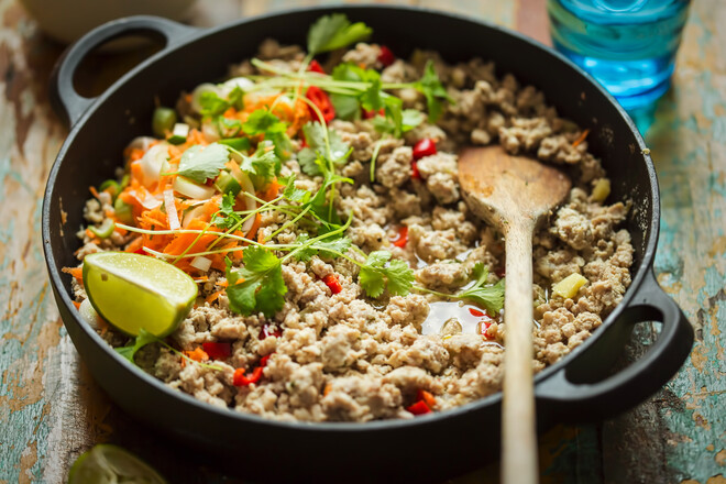 A black skillet holds cooked ground meat with vegetables like carrots and cilantro, alongside lime wedges. A wooden spoon rests in the pan on a rustic wooden table.