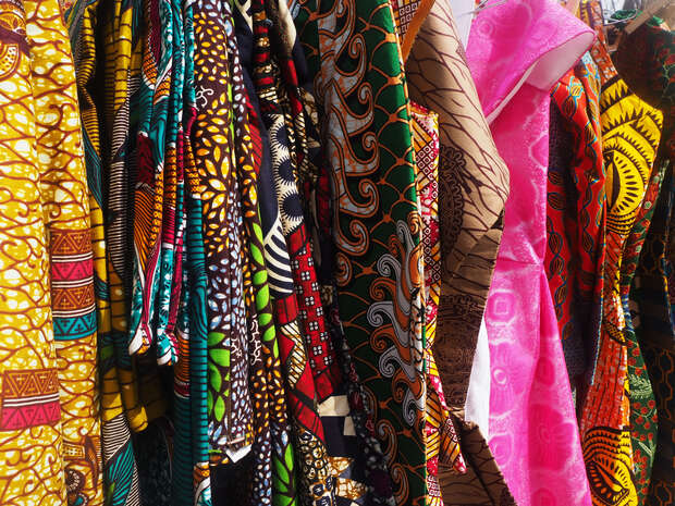 Fabrics hang closely together, displaying vibrant patterns and colors, including yellows, greens, and pinks. They are arranged in a row, suggesting a lively market or fabric store setting.