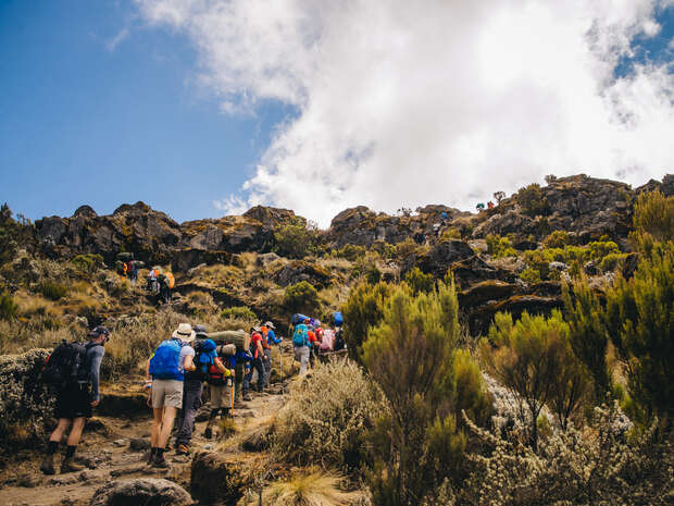 Hikers ascend a rocky trail, surrounded by shrubs and grassland. The sky is partly cloudy, and mountainous terrain rises ahead, indicating a challenging climb in an outdoor adventure setting.