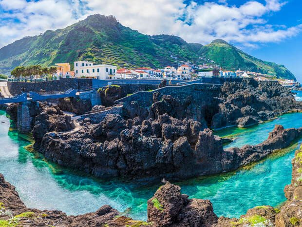 Rocky cliffs rise majestically, enclose turquoise waters, beside a coastal town. White buildings with red roofs line the shore. Verdant hills and scattered clouds complete the scenic backdrop.
