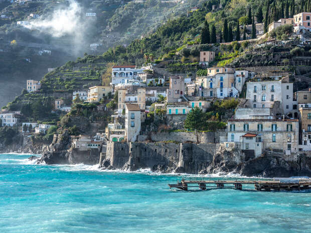 Colorful buildings line a steep hillside, perched above a rugged coastline. Blue waves crash below, while lush greenery and misty air envelop the scenic village backdrop.