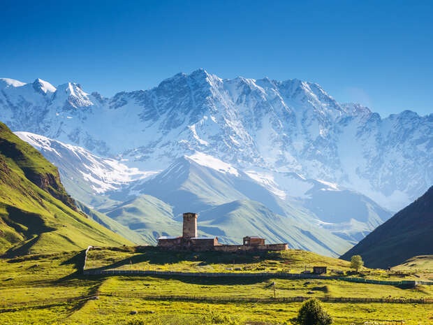 A stone tower stands in a lush green valley, surrounded by expansive grassy hills, while imposing, snow-capped mountains rise majestically in the background under a clear blue sky.