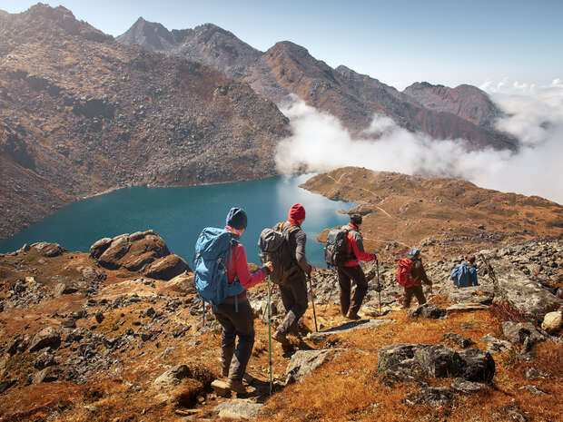 Hikers descend a rocky trail with trekking poles, wearing backpacks, overlooking a serene blue lake surrounded by mountainous terrain and scattered clouds under a bright sky.