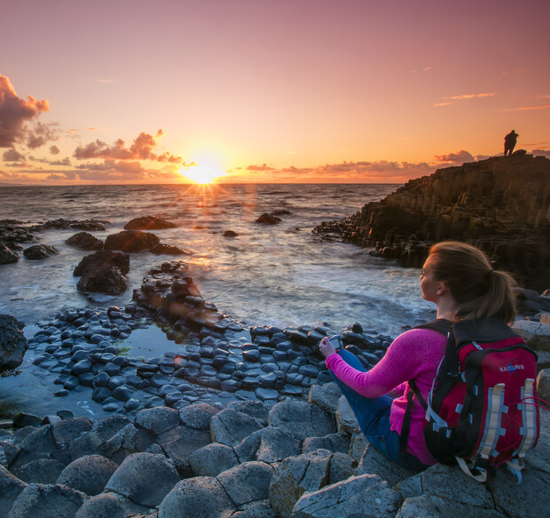 A person with a backpack sits on hexagonal rock formations, observing a vivid sunset over the ocean. Another figure stands on distant rocks, enhancing the serene coastal scene.