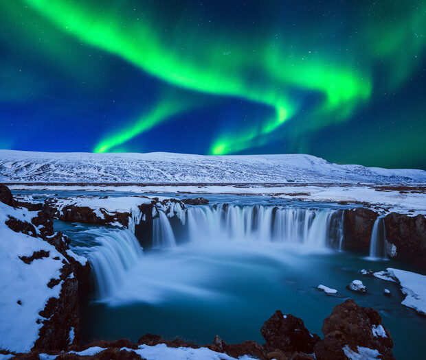 Waterfall cascading over rocks under vibrant green aurora. The snowy landscape surrounds the river, and the sky is deep blue with stars, creating a serene, magical atmosphere.