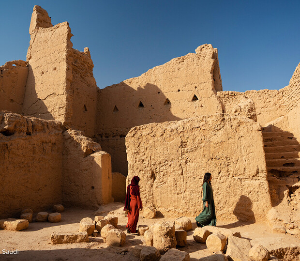 Two individuals in traditional attire walk among ancient ruins featuring beige stone walls and scattered rocks under a clear blue sky. Small triangle openings appear in the ruins. Text: "Saudi".
