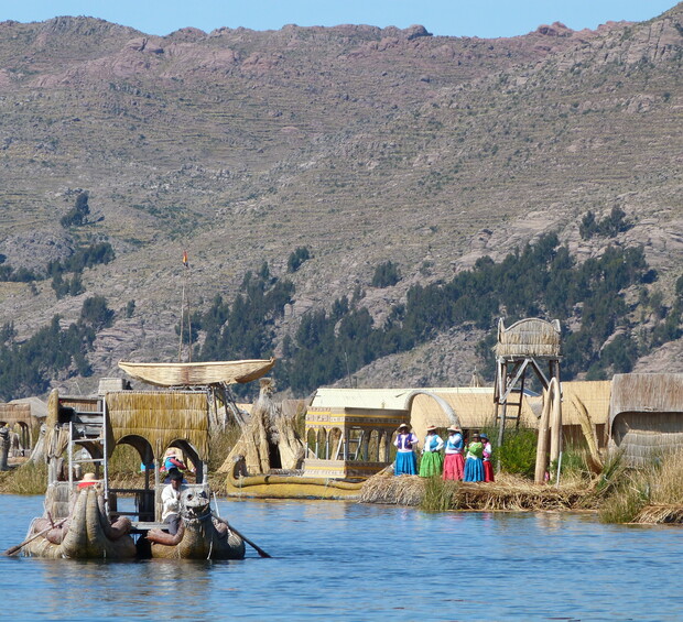 A reed boat floats on a lake near a village made of reed structures. In the background, people in colorful clothing and mountainous terrain enhance the scene's cultural and natural setting.