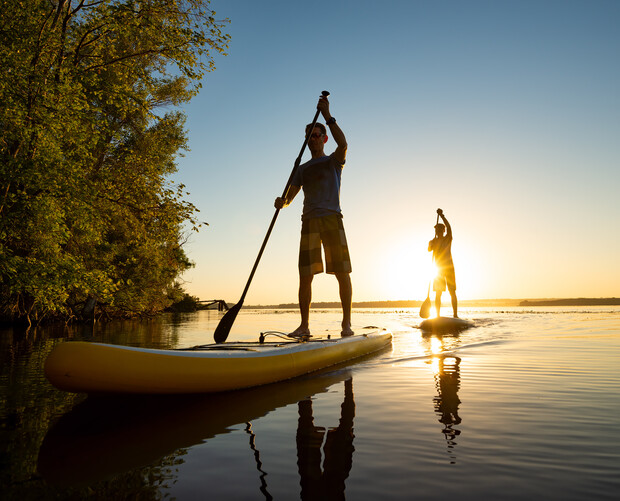 Two people are paddleboarding on calm water during sunset. The foreground paddler is silhouetted against the sun, with trees lining the shore to the left.