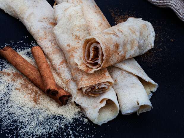 Rolled crepes sprinkled with cinnamon lie on a dark surface alongside two cinnamon sticks and scattered sugar, suggesting a sweet, aromatic dessert setting.