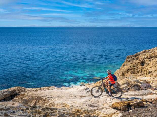 A cyclist stands on rocky terrain beside a vast blue ocean, wearing a helmet and backpack, facing the water under a clear, lightly clouded sky.