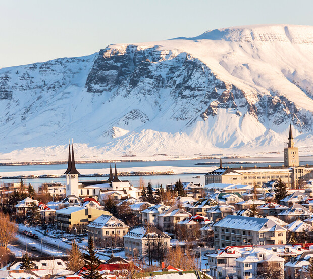 Snow-covered townscape with church spires standing still against a backdrop of a vast mountain range, under a clear sky, creating a serene, wintry atmosphere.