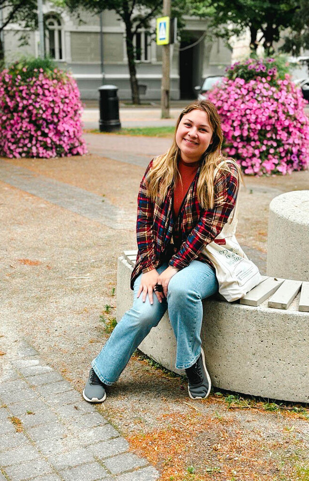 A person sits on a round concrete bench in a park, smiling, wearing a plaid jacket and jeans. Behind them are pink flower bushes and a building in the background.