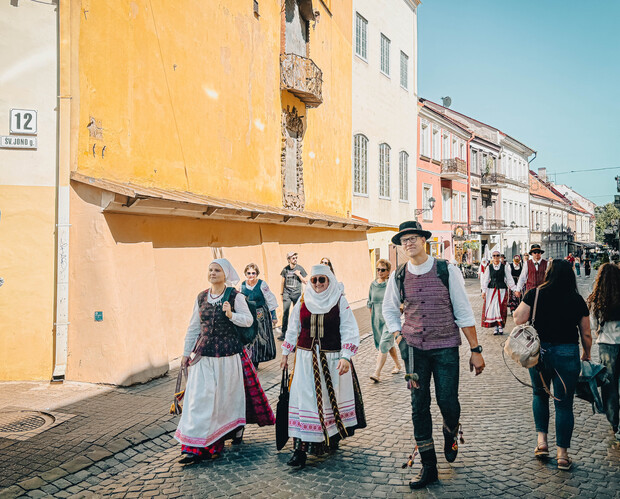 People in traditional attire walk along a cobblestone street beside old, colorful buildings. Some are wearing hats and carrying bags, and others follow. The sunny day illuminates the vibrant scene.