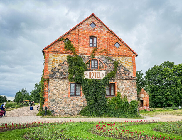 A rustic brick and stone building features a sign reading "HOTEL," with ivy climbing the walls. It stands amid grassy landscaping with a cloudy sky and trees in the background.