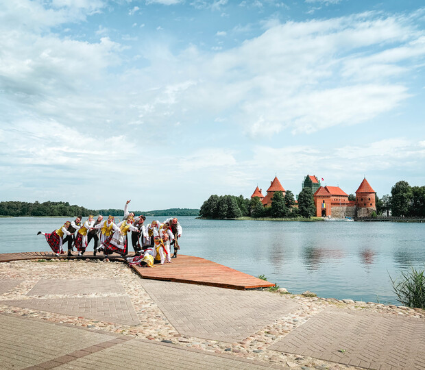 People in traditional costumes dance and pose on a lakeside platform, with a red-roofed castle on an island and lush greenery under a partly cloudy sky.