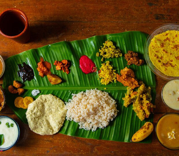 A banana leaf serves as a plate, holding rice, various curries, chutneys, and papadum. Accompanying dishes and a glass cup are arranged on a wooden table background.