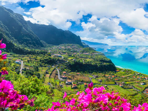 Purple flowers bloom in the foreground overlooking a lush valley with terraced fields, small houses, and distant mountains under a partly cloudy sky by a turquoise sea.