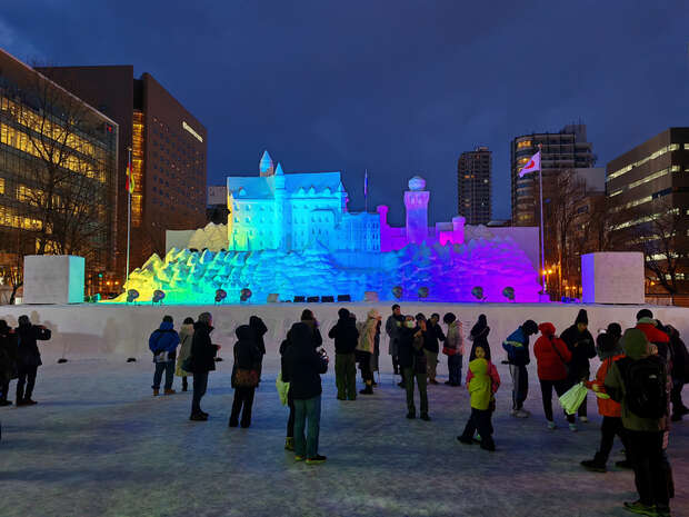 A large snow sculpture resembling a castle is illuminated with colorful lights. People stand around observing, surrounded by tall buildings during night in an urban setting.