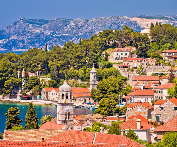 Coastal town nestled among lush greenery and red-roofed buildings, featuring a stone clock tower by the water. Mountains rise in the background under a clear blue sky.