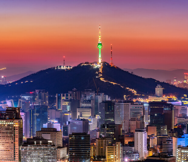 A brightly lit tower stands on a hill, overlooking a bustling cityscape filled with skyscrapers during sunset, with a gradient sky transitioning from orange to deep blue.